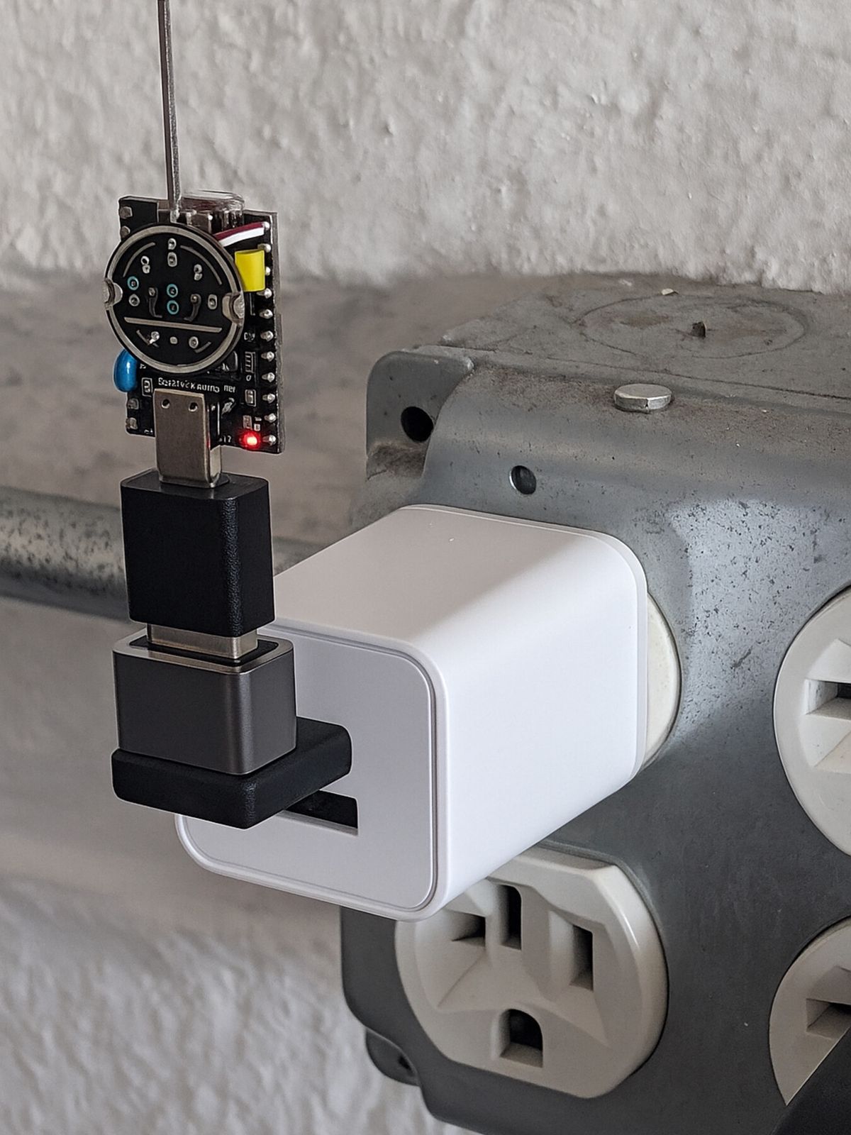 An ESP32 board with a welding-wire whip antenna plugged into a small USB-power adapter, which is plugged into a wall outlet beside an industrial junction box