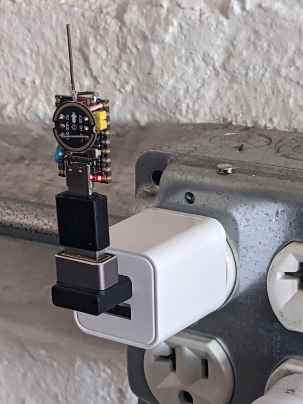 An ESP32 board with a welding-wire whip antenna plugged into a small USB-power adapter, which is plugged into a wall outlet beside an industrial junction box