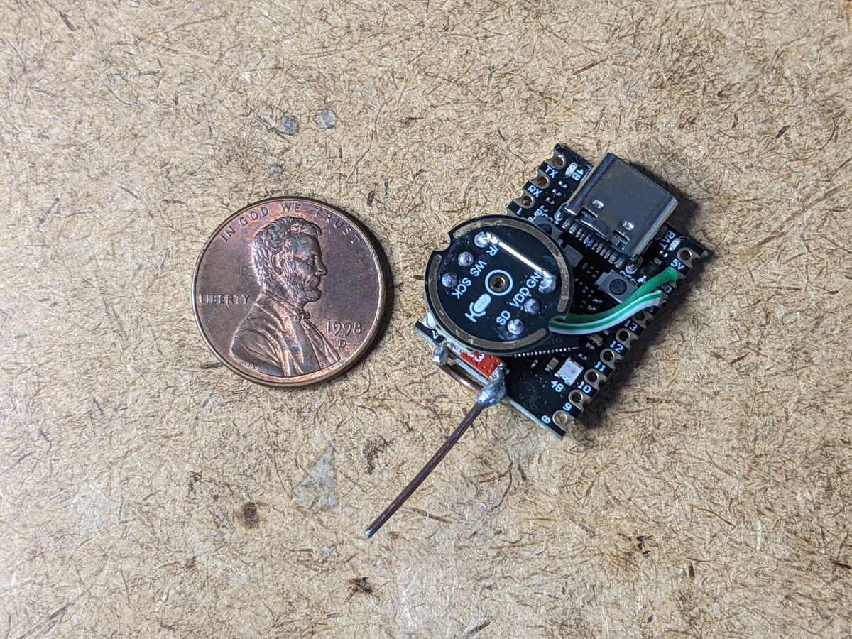 An ESP32-S3 dev board on a wooden workbench, with a thin piece of welding wire soldered to it as a vertical whip antenna, and a US penny placed beside it for scale